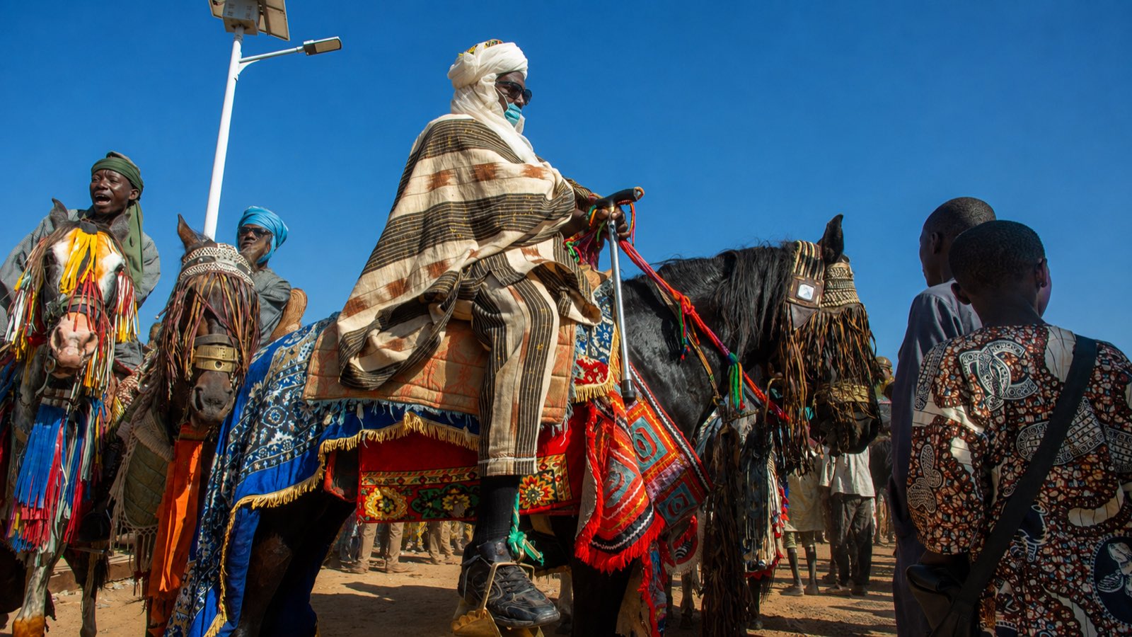 Cavaliers en boubous traditionnels sur leurs chevaux harnachés de tissus colorés.